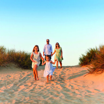 Happy family of four, young parents and little kids, enjoying summer vacation in nature, taking walk on sandy beach, holding each others hands, girls sisters running ahead while parents walking behind