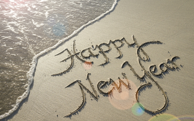 Happy New Year message handwritten in smooth sand with sunrise lens flare over oncoming wave on the beach