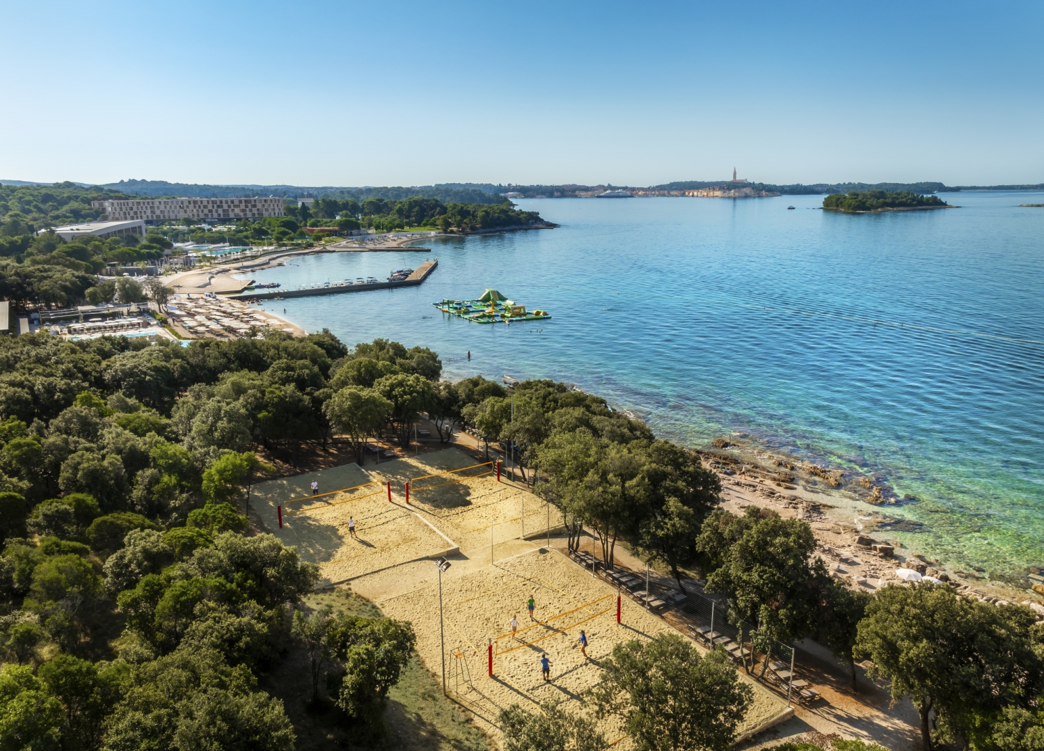 Luftaufnahme von Resort am Meer mit Strand und Volleyballplatz, umgeben von Bäumen.